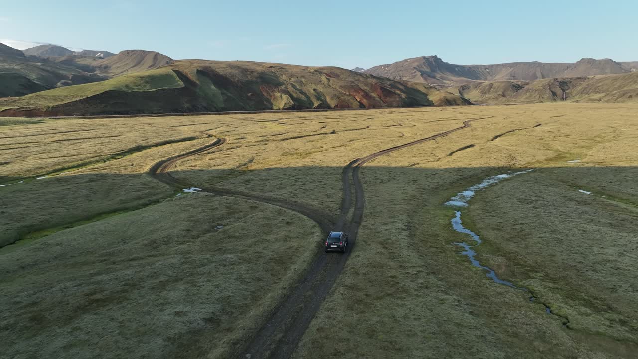 A lone off-road vehicle travels through a vast, remote valley in northern Iceland, surrounded by rugged mountains and winding streams under a clear sky.