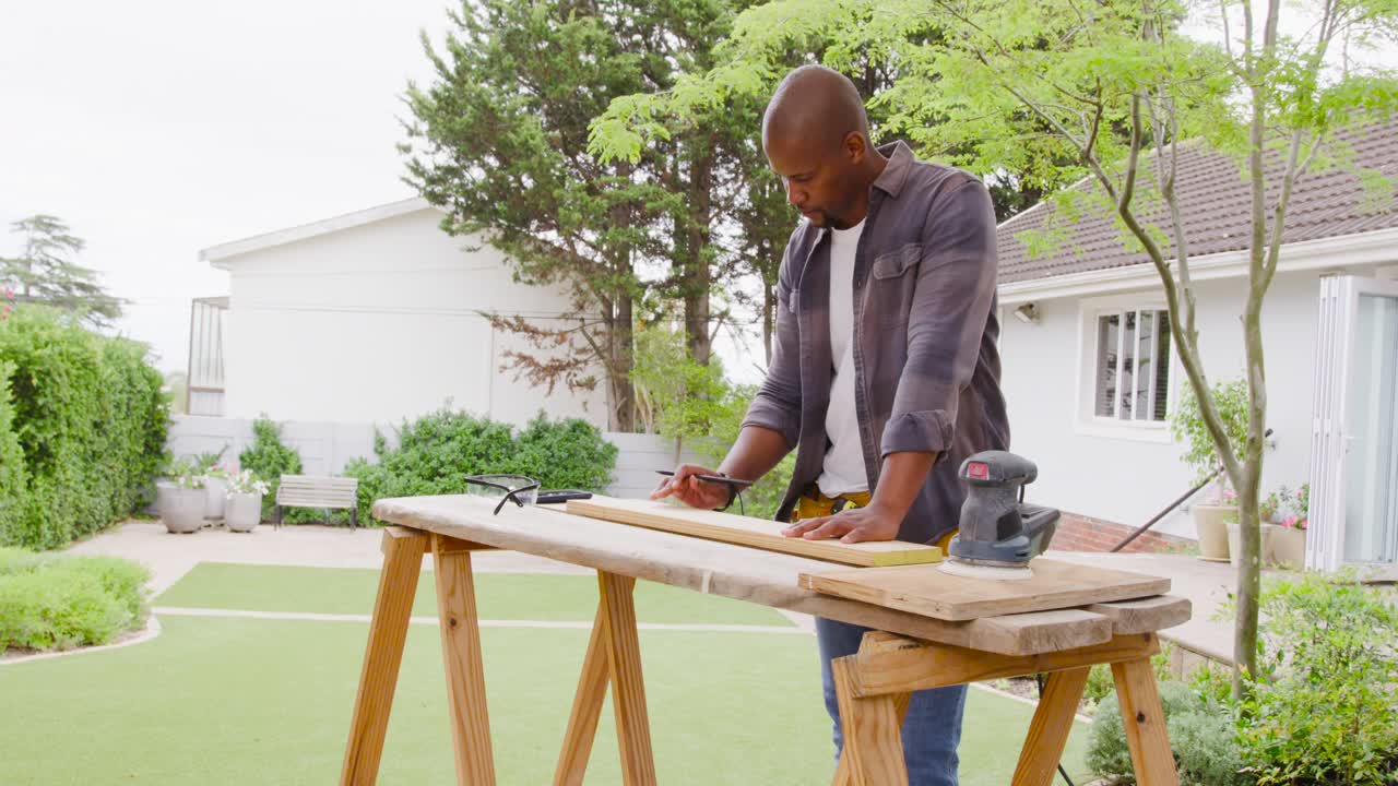Homme afro-américain en chemise à carreaux mesurant et marquant du bois d'œuvre avec un crayon sur des tréteaux pour la coupe.