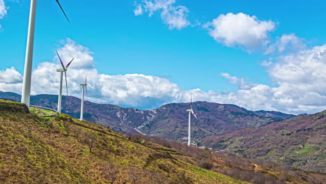 static view of white wind turbines moving in timelapse over a hilly terrain with white clouds passing by on a bright sunny day