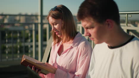 close-up of young woman smiling while reading a book and showing it to her friend outdoors, the man is focused on her gesture, blurred background features greenery and urban setting