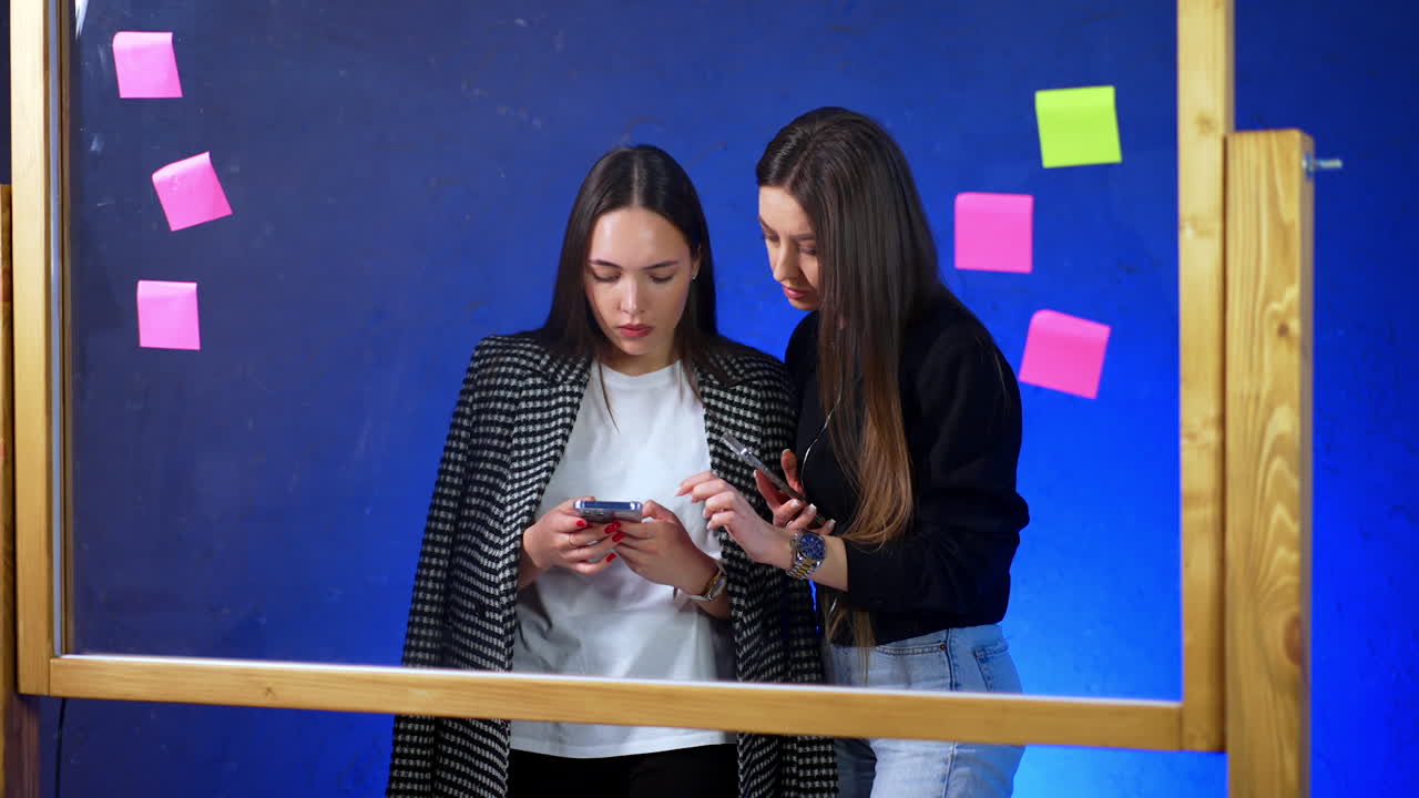 Caucasian women stand at the glass board with stickers. Brunette ladies both hold the phones but are focused on the phone of the girl in jacket.