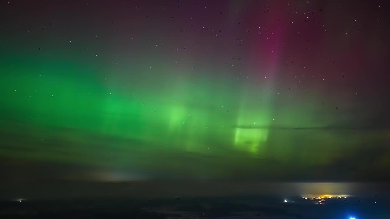 Un'aurora boreale di un verde vivido si snoda nel cielo notturno stellato sopra le luci di una città lontana in un timelapse orizzontale