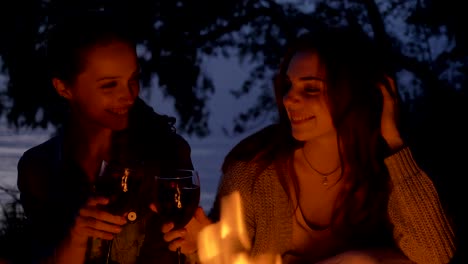 dos jóvenes caucásicas se sientan por la noche junto al fuego en la naturaleza, beben vino, sonriendo