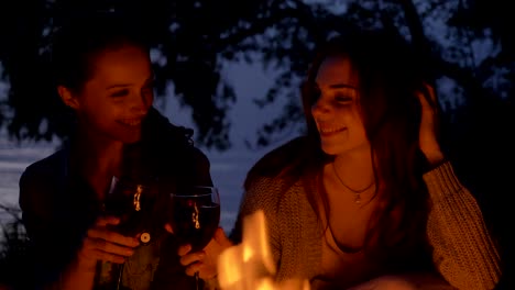 two young caucasian girls sit in the evening by the fire in nature, drink wine, smiling