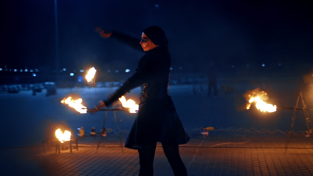 Woman in spooky costume dances gracefully holding a string with burning ball. Fire show performance at night.
