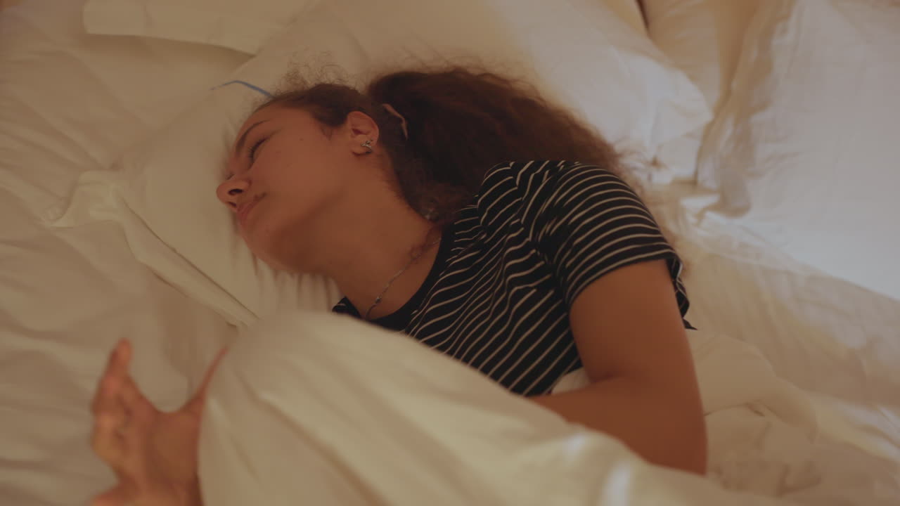 Fredfyldt kvinde ligger med pude. Ung kvinde i stribet tøj læner sig forsigtigt tilbage med et roligt udtryk. Kvinde virker rolig og afslappet, mens hun ligger komfortabelt med blød belysning og pude.