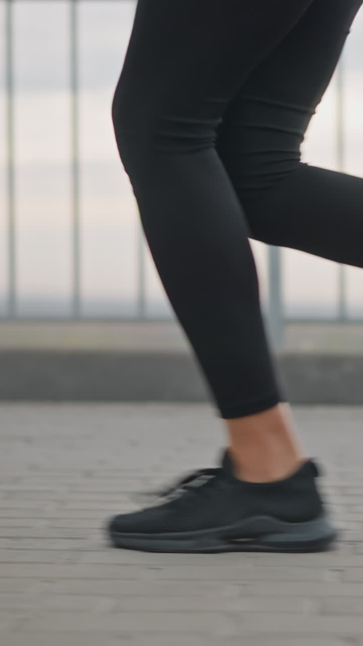Close-up view of woman's legs in black leggings and black sneakers jogging along a paved track, iron fence and distant view of a river add dynamic outdoor fitness ambiance