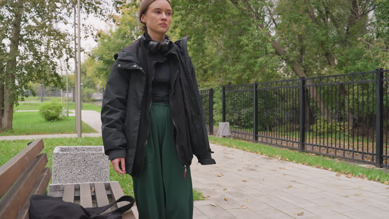 white woman sitting on bench, pensive expression, black jacket and green pants, headphones around neck, backpack on bench, overcast autumn park with trees and path, slow walk then seated rest, varied