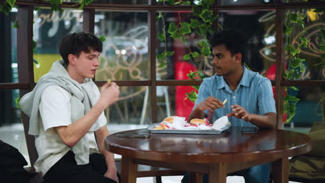 Two colleagues sitting adjacent to each other while sharing burger tray on table, having friendly conversation during break casual lunch meeting, discussing way forward and exchanging ideas