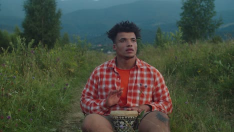 handsome hipster african man playing djembe drum on mountain hill at sundown