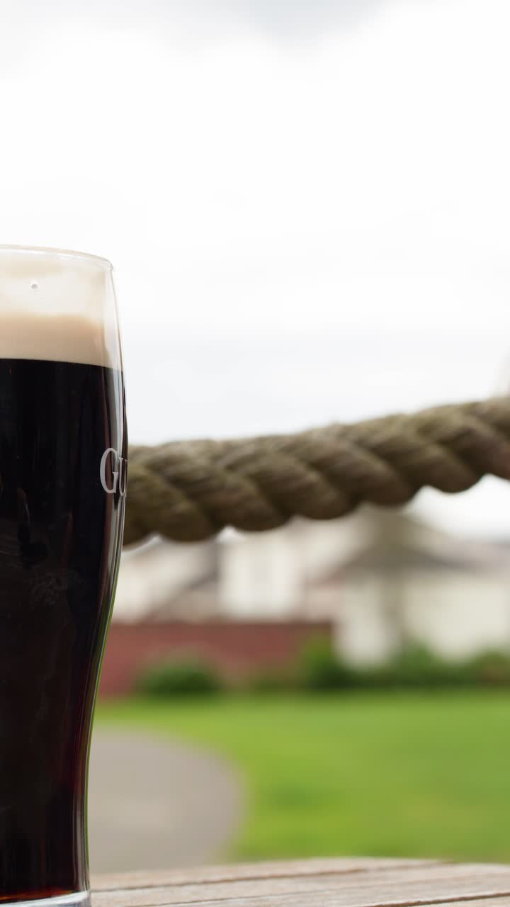 Hände stoßen dunkle Biergläser draußen auf einem Holztisch an, natürliches Tageslicht, geringe Tiefenschärfe.
