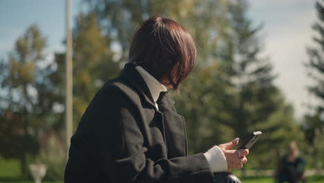 lady seated outdoors looking thoughtfully, focused on phone in hand, ring on finger, with a blurred background of greenery and trees on a sunny day