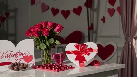 romantic valentine's day setup with red roses, heart shaped gift box, greeting card, and chocolates arranged on white table, illuminated by soft daylight through window