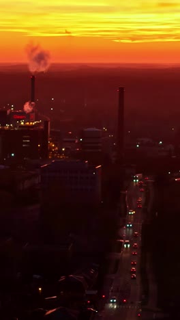 an atmospheric aerial shot of jelgava, latvia, captures a factory smokestack emitting a plume of smoke against a vibrant orange sunset sky, with city traffic visible on the streets below - vertical