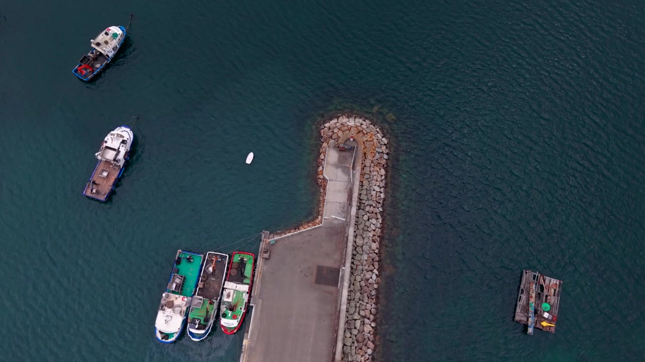 Dronebillede ovenfra af fiskebåde fortøjet langs en stenmole og bølgebryder i en rolig grøn havn på Galiciens kyst i Spanien