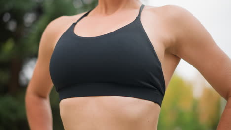 Close view of a woman in black sportswear pushing her chest out, showcasing confidence and strength, with hands on her waist, outdoors in a serene environment