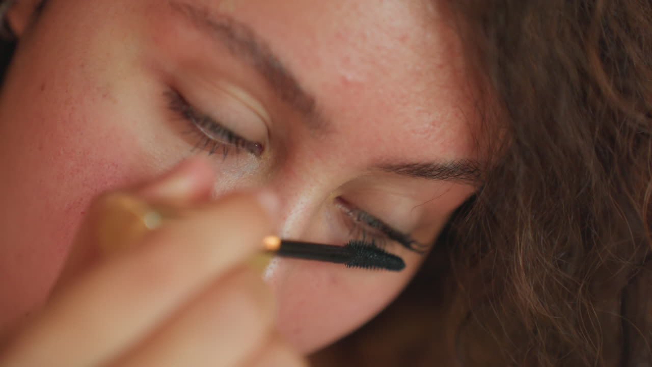 Hotel room closeup multiracial woman applying mascara, soft natural light, dewy skin, steady hand moving wand along upper lashes, focused grooming ritual, intimate calm mood, fine lash detail
