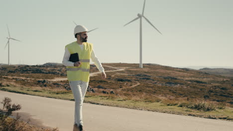 ensuring a sustainable future for our planet, a professional engineer in a white helmet and reflective vest uses a tablet to audit wind turbines in a field of clean energy generators on a sunny day