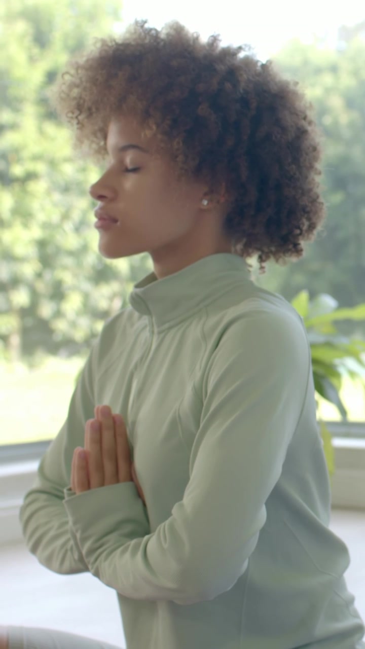 Vertical video: Meditating woman in green top starting with hands pressed at chest by window, plant