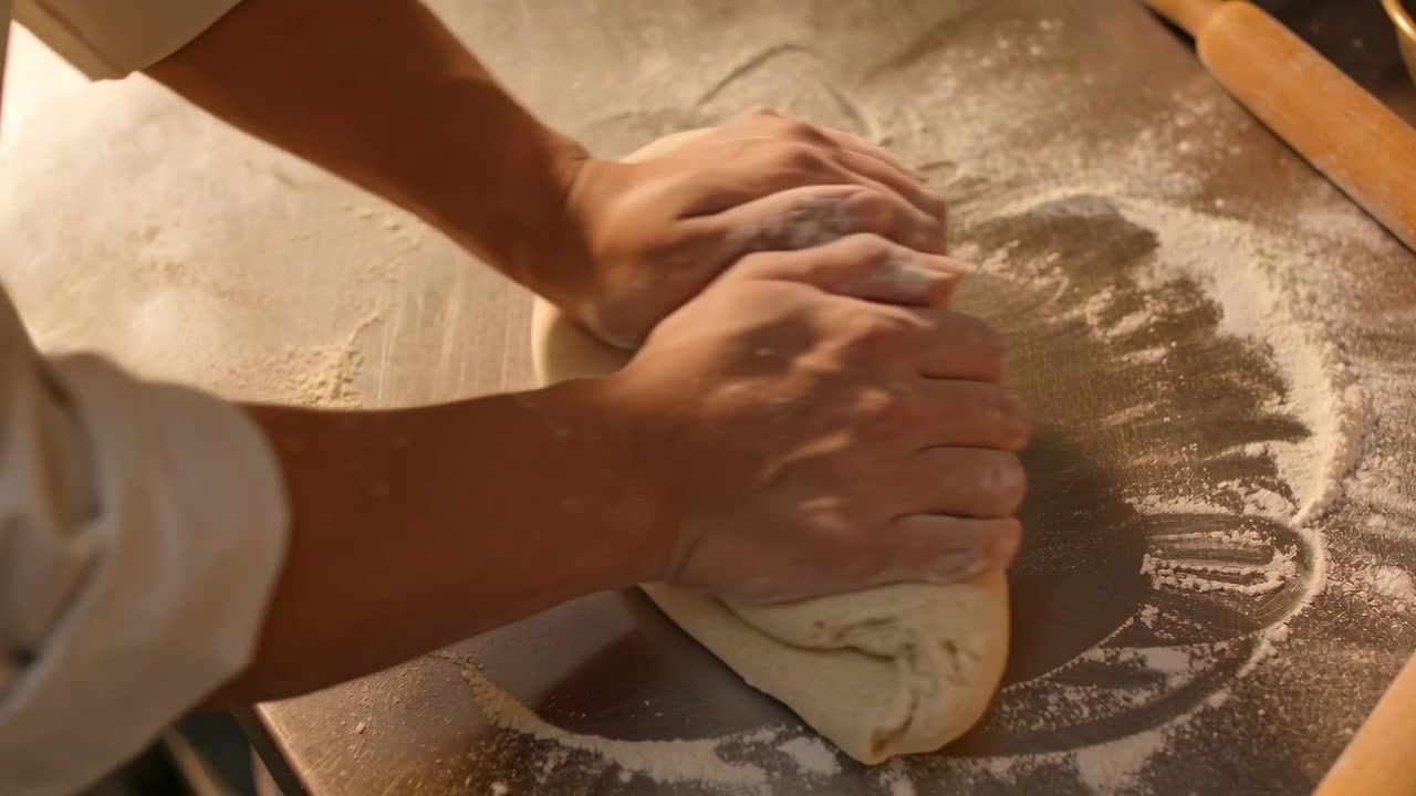 Fornaio con le maniche arrotolate che impasta, piega e modella l'impasto su un bancone di metallo, con un mattarello.