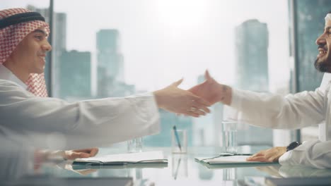 two successful emirati businessmen in white traditional outfit sitting in office and signing contract. business partners shake hands on lucrative investment. saudi, emirati, arab businessman concept.