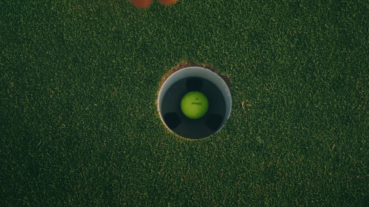 Top-down slow motion shot of a yellow golf ball rolling into the cup, bouncing inside the hole, while a male golfer’s arm reaches in to retrieve it. A close, dynamic moment capturing golf precision