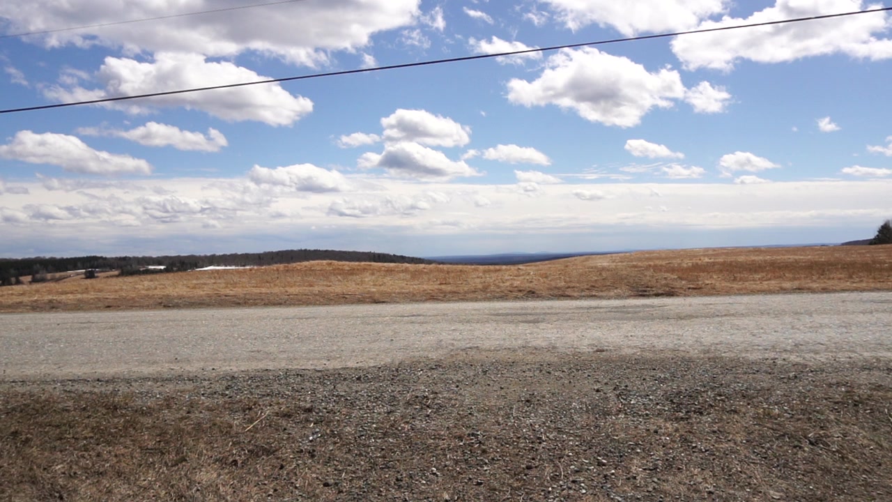 Eye-level, panoramic view of a gravel road bordering a vast, dormant brown agricultural field. Distant rolling hills under a vibrant blue sky with scattered white cumulus clouds. Power line across top