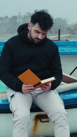 man sitting on a boat on the beach finishes reading a book on a gloomy day