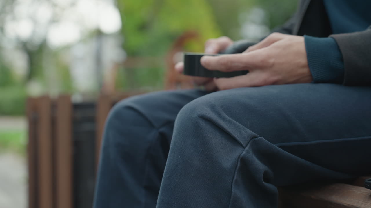 Mand tjekker telefon, person hviler sig på bænk og kigger på skærm, person slapper af på parkbænk og bruger mobilenhed, person sidder på parkbænk og er fokuseret på sin smartphone-skærm