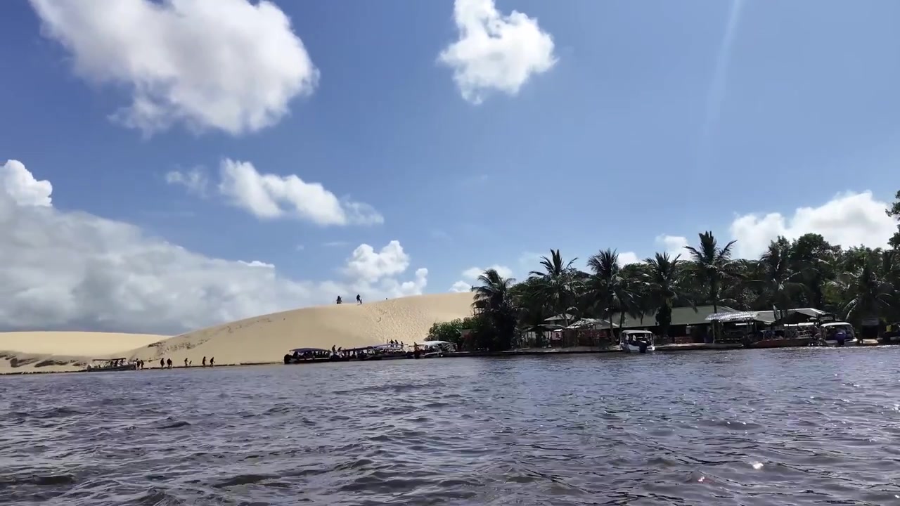 Toma amplia a nivel del agua moviéndose a lo largo del río Preguiça, capturando la parada del pueblo turístico en Vassouras, rodeado de palmeras, dunas de arena blanca y barcos turísticos atracados a lo largo de la orilla en Maranhão