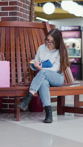 joven sentada en un moderno banco de madera en un centro comercial bien iluminado, con las piernas cruzadas, absorta en un libro con la luz reflejada en las baldosas brillantes del centro comercial y las bolsas de compras a su lado