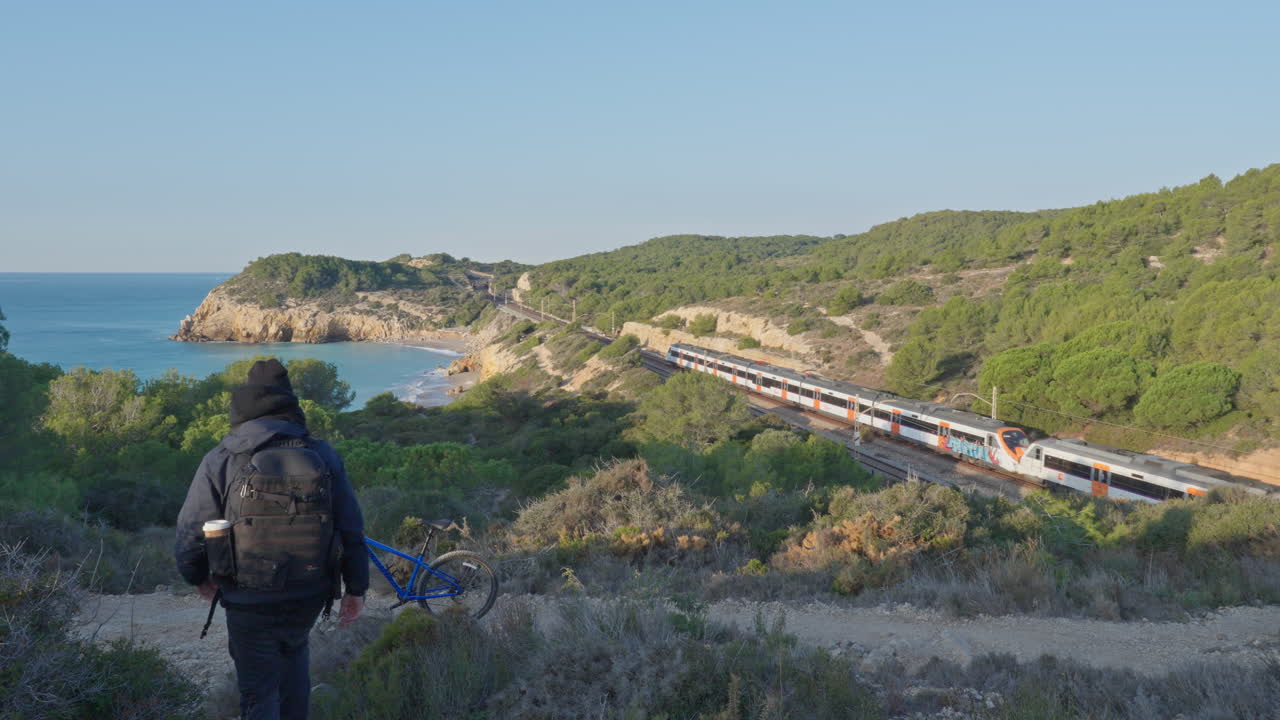 paisaje con el mar lejos, tren a distancia saliendo y ciclista camina hacia su bicicleta dejando la soleada escena montañosa