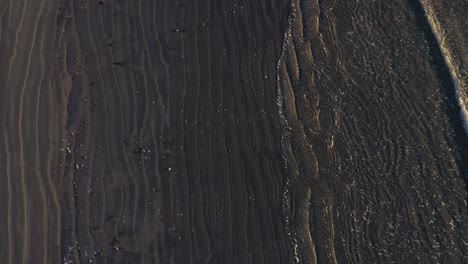 aerial top down slowly flying over a wave coming in on a grey sand beach with cool patterns in the sand and surf