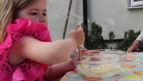 medio disparo de una chica joven en un vestido de princesa rosa, agitando un plato de mezcla de pastel como un padre agrega una cucharada de azúcar