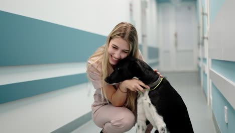 Portrait-of-a-happy-blonde-girl-with-her-black-and-white-dog-in-a-blue-corridor-in-a-veterinary-clinic.-Happy-blonde-girl-with-her-dog-posing-in-a-veterinary-clinic