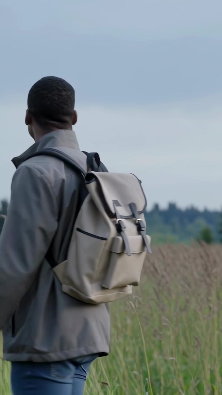 Vertical video: Turning man in gray jacket jeans walking in grass toward trees backpack, copy space