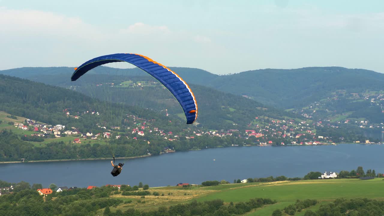 un solo parapente hace un largo giro a la derecha contra montañas boscosas con pequeños edificios y un gran lago