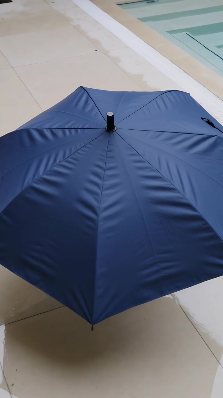 Video verticale: Un ombrello pieghevole blu navy che si apre a scatto, con la sua cupola che si espande sulle piastrelle a bordo piscina.
