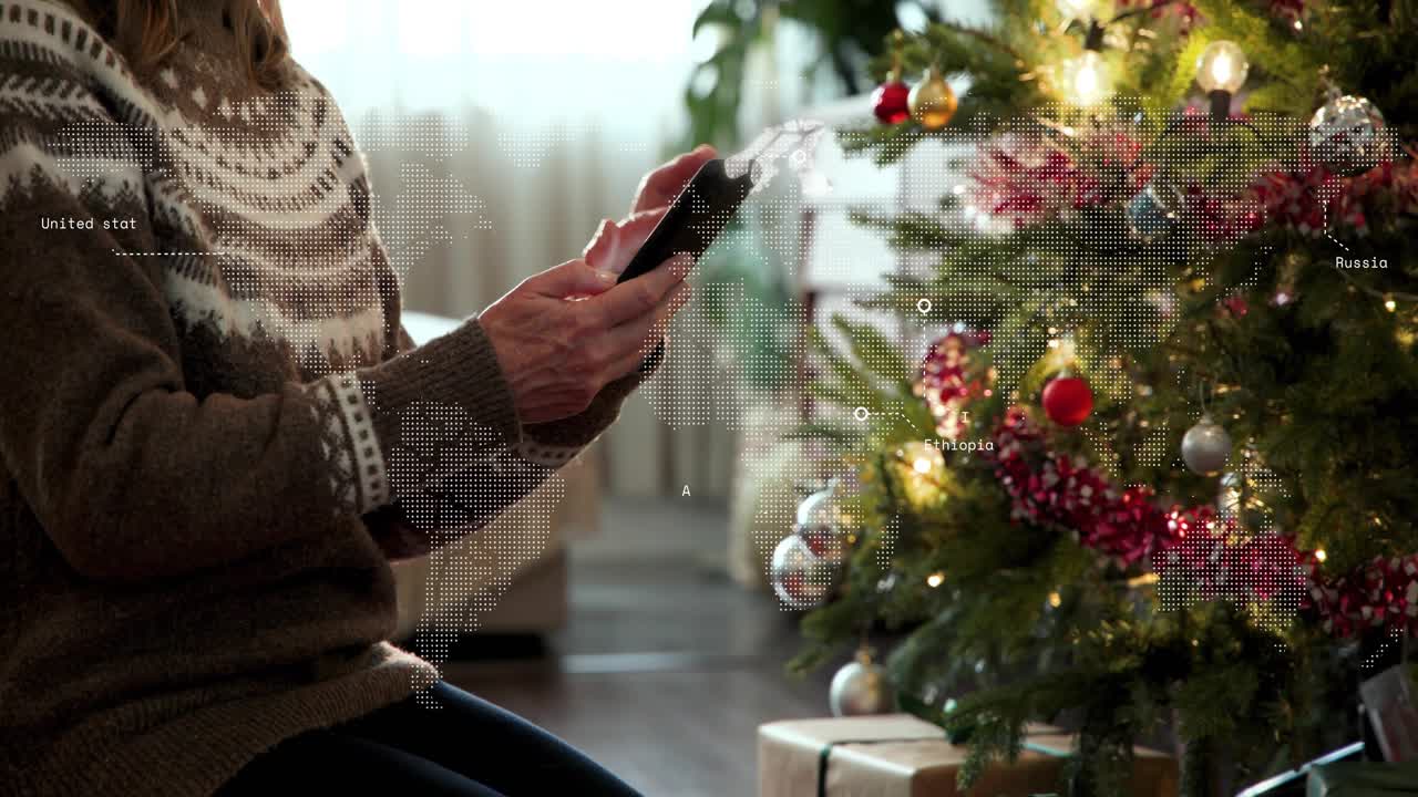 Mature woman kneeling by Xmas tree tapping phone initiating e-commerce tracking world-map overlay