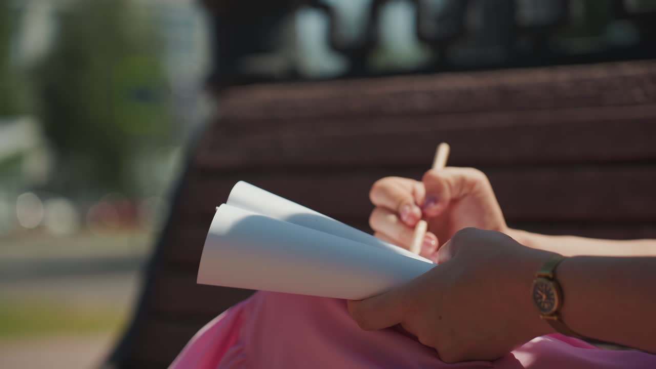 Primo piano di mani che scrivono su un quaderno su una panchina, donna con orologio che sfoglia le pagine mentre batte con una matita, sfondo urbano sfocato con bokeh di passanti, luce solare calda, atmosfera rilassata pomeridiana, spontaneo