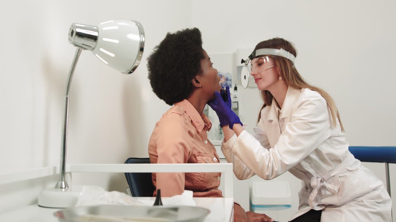 Giovane dottoressa europea otorinolaringoiatra esamina la gola di una donna afroamericana con un divaricatore in legno