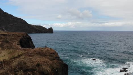 The-drone-captures-a-majestic-rock-formation-on-Madeira-Island,-where-two-figures-stand,-dwarfed-by-the-rugged-landscape,-highlighting-the-grandeur-of-nature