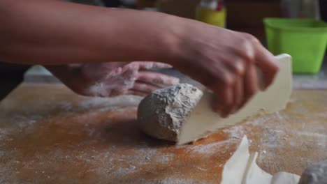 ball of freshly risen dough being shaped and shuffled by hand and scraper tool on wooden kitchen tabletop, filmed as closeup slow motion shot