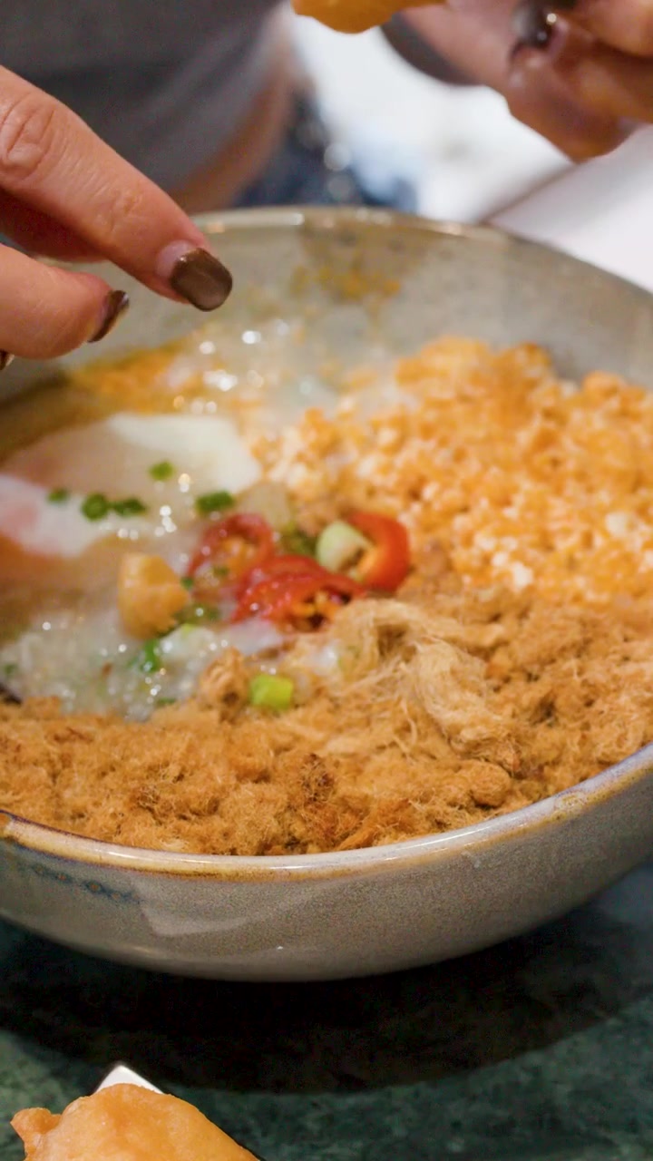 Close-up tangan merobek cakwe ke dalam semangkuk bubur nasi gurih Asia.