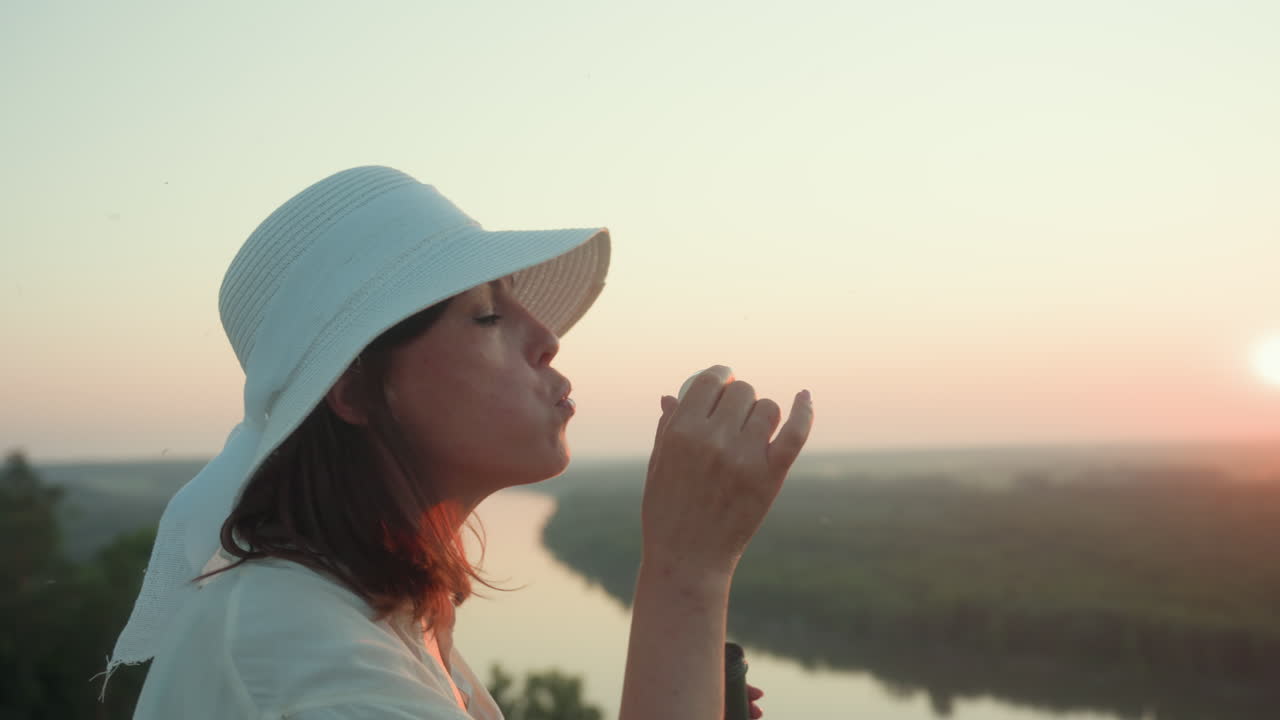 Side view of lady wearing white sun hat gently blowing soap bubble toward glowing sunset sky, with winding river valley and grassy hillside in soft golden light