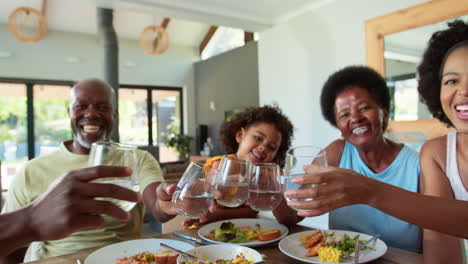 Mehrgenerationenfamilie-Sitzt-Zu-Hause-Am-Tisch-Und-Jubelt-Mit-Wasser-In-Richtung-Kamera