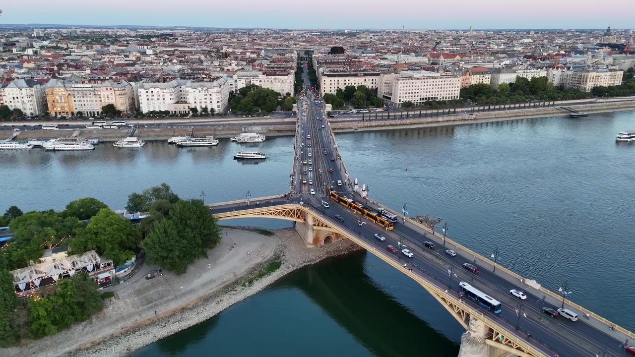 Budapest, Ungheria, il Ponte Margherita che attraversa il Danubio accanto all'Isola Margherita al tramonto, con tram, barche e traffico serale nel paesaggio urbano sul lungofiume, vista aerea