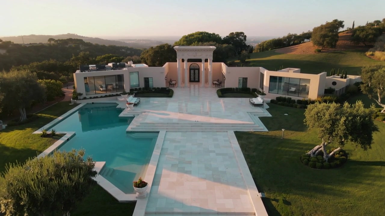 Una telecamera aerea si avvicina rivelando una villa moderna in campagna con piscina a forma di croce e passerella.
