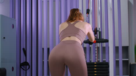 rear view of young woman in brown activewear performing cable weight pull in gym, engaging core and back muscles, focusing on strength training, fitness routine, and body toning