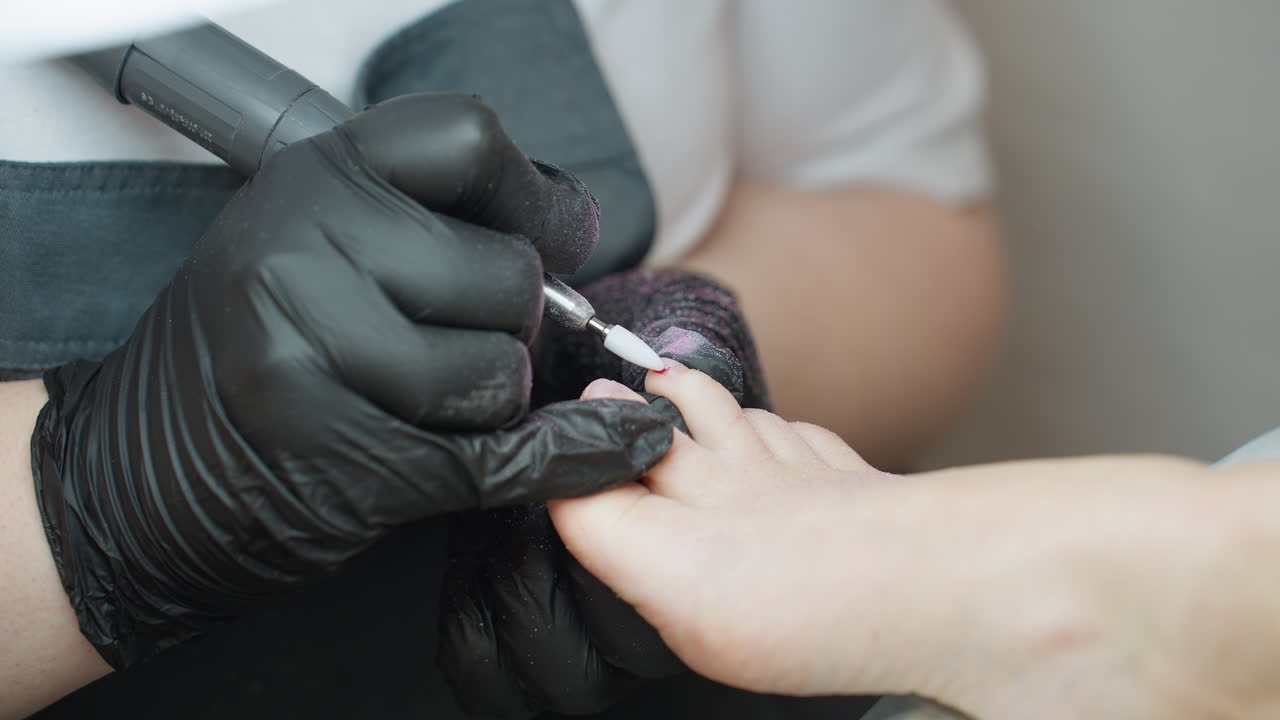 Close up view of nail technician wearing black gloves using electric file on client toe during pedicure procedure, with focus on precision and nail care in professional salon environment